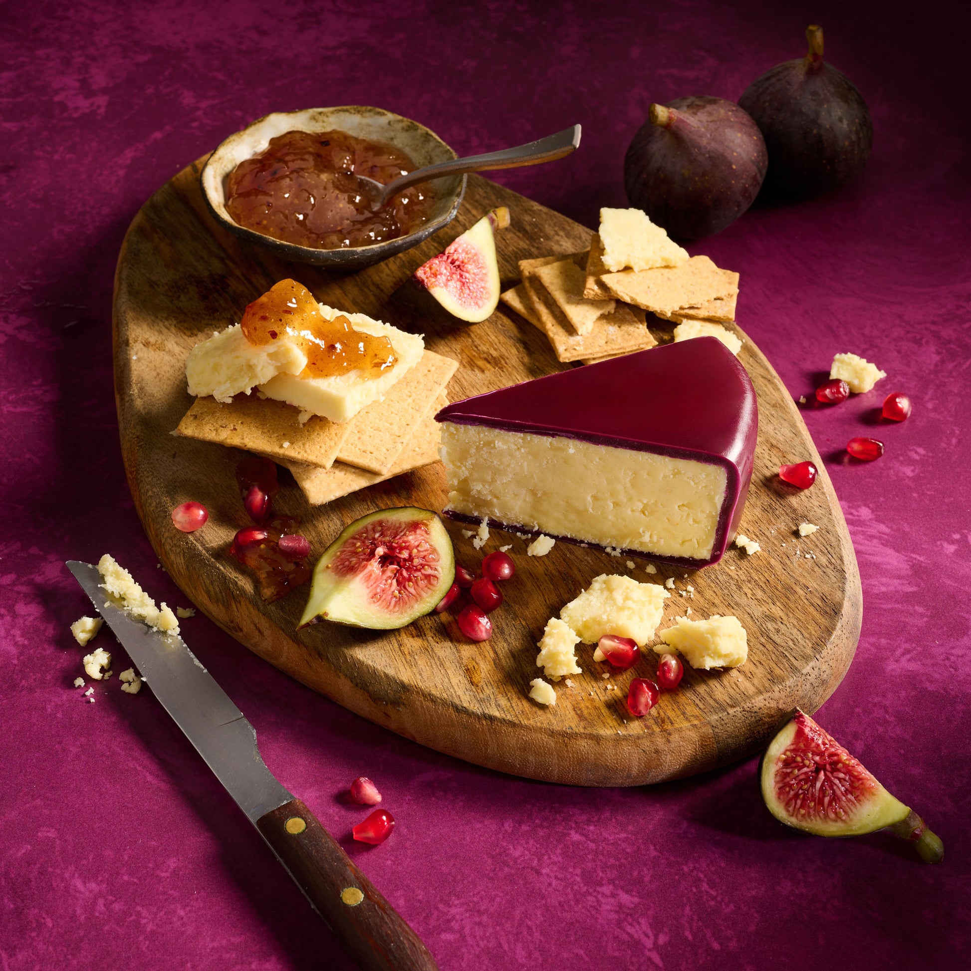 Wooden cutting board with cheese, figs, and crackers on a purple background